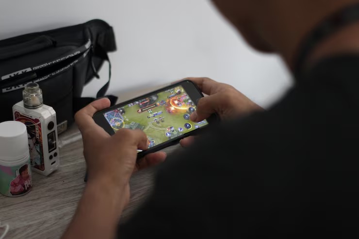 Person playing a mobile video game on a smartphone, holding the device horizontally at a desk with personal items beside them.