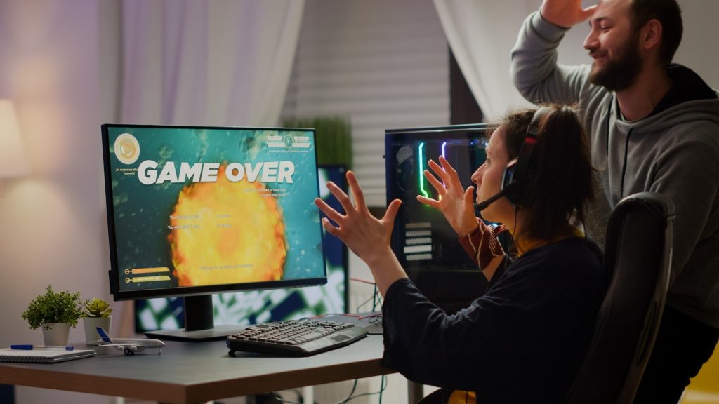 Frustrated female gamer wearing headset reacting to ‘Game Over’ screen while male friend celebrates behind her at gaming setup