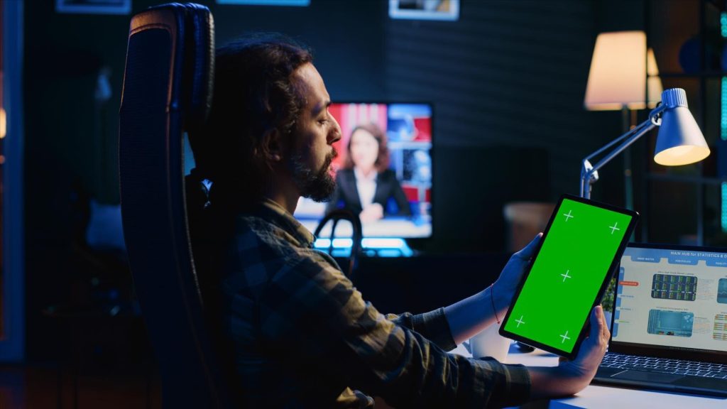 Man holding tablet with green screen in a dark home office.